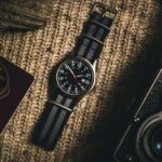 Klassiek polshorloge met zwart NATO-bandje, gefotografeerd op een wollen ondergrond met vintage camera.