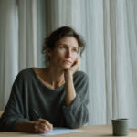 Vrouw zit peinzend aan een houten tafel bij het raam met een pen in de hand tijdens het invullen van een vragenlijst over haar gezondheidsdoelen